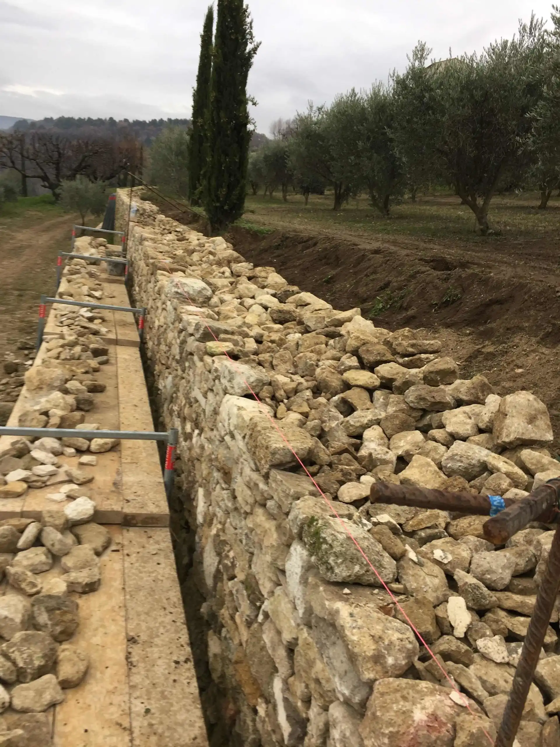 Construction d’un mur de soutènement en pierre naturelle dans une oliveraie avec remplissage drainant en pierres.